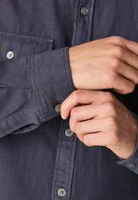 Dunkelgraues Hemd mit Knopfleiste, Brusttasche und strukturiertem Stoff. Es hat graue Knöpfe und verstellbare Manschetten und hebt einfache Designmerkmale hervor.