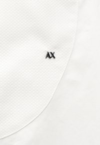 Gros plan d’un tissu blanc texturé avec un petit logo noir « AX » centré sur la surface.