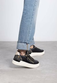 Zapatillas de cuero negro con detalles de cremallera y acentos beige, combinadas con vaqueros de mezclilla claros con dobladillo. Presentan una suela blanca y patrones texturizados.