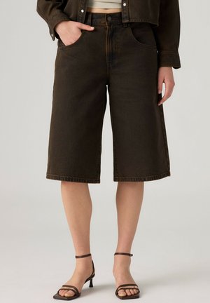 Personne portant un short en jean marron foncé au genou, une veste courte et des sandales marron à talons hauts et à brides sur un fond uni.