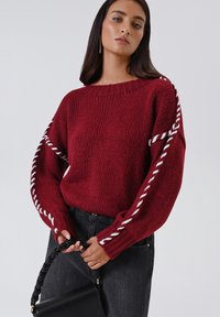 Donna che indossa un maglione rosso a maglia con dettagli bianchi attorcigliati sulle maniche, mentre tiene una borsa nera con manico intrecciato, su uno sfondo semplice.