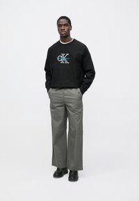 Μαύρο φούτερ με λογότυπο «CK» σε μπλε χρώμα, σε συνδυασμό με γκρίζο, φαρδύ παντελόνι και μαύρα παπούτσια. Απλό φόντο για έμφαση.