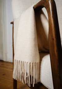 Cremefarbene, wollene Decke mit Fransen, drapiert über einen dunkelholzigen Sessel mit beiger Polsterung auf einem Holzboden.