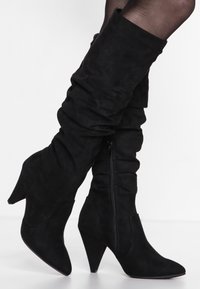 Botas negras de ante, hasta la rodilla y con un diseño flexible. Cuentan con un tacón afilado y una cremallera lateral para ponérselas fácilmente.