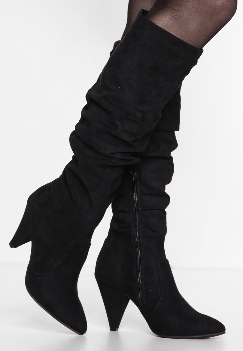 Botas negras de ante, hasta la rodilla y con un diseño flexible. Cuentan con un tacón afilado y una cremallera lateral para ponérselas fácilmente.