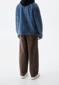 Persona con chaqueta de mezclilla azul, pantalones marrones sueltos con bolsillos traseros y zapatillas blancas, de pie frente a un fondo blanco.