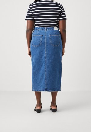 Frau trägt ein gestreiftes kurzärmeliges Shirt, einen blauen hoch taillierten Midi-Denimrock und schwarze Sandalen mit Absatz, stehend vor einem einfarbigen Hintergrund.