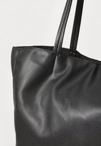 Črna torba iz teksturiranega sintetičnega materiala z dvema dolgima ročajema in mehkimi, gladkimi površinami. Oblikovanje se ponaša s preprosto, neokrašeno obliko.