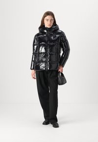 Chaqueta acolchada negra de brillo alto con capucha, cremallera frontal y patrón de acolchado horizontal. Combinada con pantalones de pierna ancha y un bolso pequeño.