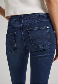 Dark blue denim jeans with a slim fit, featuring two back pockets and visible stitching detail. The fabric appears smooth with slight texture.