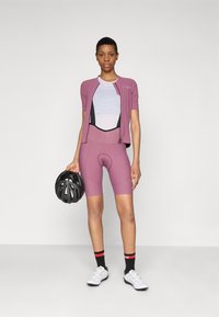Cyklistický outfit v svetlofialovej farbe. Krátky rukáv s prešívanými detailmi, vystužené kraťasy, čierna prilba a biele cyklistické topánky s akcentmi.