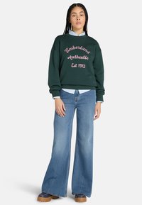 Tummanvihreä huppari, jossa on vaaleanpunaisella käsinkirjoitettu teksti "Timberland Authentic Est 1973". Yhdistetty vaaleansinisisiin leveälahkeisiin farkkuihin ja ruskeisiin platform-bootseihin.