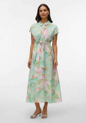 Mujer con un vestido midi floral de tonos pastel, con cuello, mangas cortas, cinturón anudado en la cintura y sandalias de tacón de color claro.