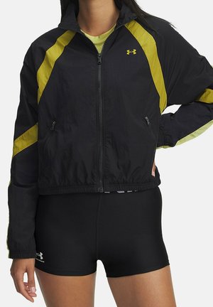 Person trägt eine schwarz-gelbe Jacke mit Reißverschluss über einem grünen Shirt und schwarzen enganliegenden Shorts, steht mit einer Hand in der Hüfte.