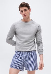 Jeune homme portant une chemise à manches longues gris clair et un short à carreaux bleu et blanc, debout devant un fond blanc uni.