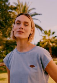 Νεαρή γυναίκα με κοντά ξανθά μαλλιά που φοράει ανοιχτό γαλάζιο αμάνικο τοπ, στέκεται σε εξωτερικό χώρο με φοινικόδεντρα στο φόντο κατά το ηλιοβασίλεμα.