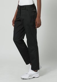 Svarta träningbyxor med en slät, matt finish, som har fickor med dragkedja och en diskret logotyp på sidan. Matchas med vita träningsskor.