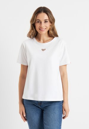 Jonge vrouw die een wit T-shirt draagt met een klein zilveren hartontwerp op de borst, gecombineerd met een blauwe jeans, lachend naar de camera.