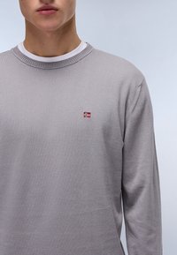 Maglione grigio chiaro a maniche lunghe con collo a costine e una piccola bandiera norvegese ricamata sul petto. Tessuto morbido e lavorato a maglia.