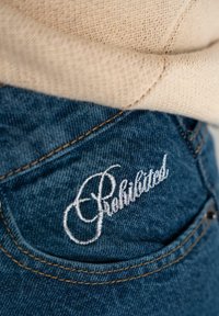 Jean en denim bleu foncé avec des coutures beiges. La poche arrière présente un texte blanc brodé "Rehabiliated". La ceinture est beige.