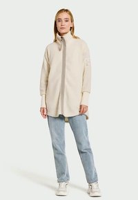 Fleece zip-up jacket in off-white, featuring a high collar, long sleeves, and a straight cut, paired with light blue jeans and white sneakers.