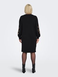 Mustast lõngast kleit pikkade varrukatega, millel on mansettidel lillelise tikandiga detailid. Kleit on lõdva lõikega, ulatudes reie keskpaigani, ja seda kantakse koos mustade pahkluu-saabastega.