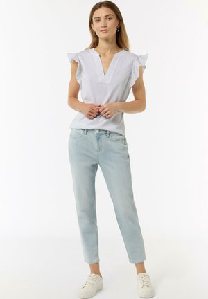 Top bianco a scollo a v con maniche a volantino, abbinato a jeans tapered azzurri chiari e sneakers bianche, in piedi su uno sfondo neutro.