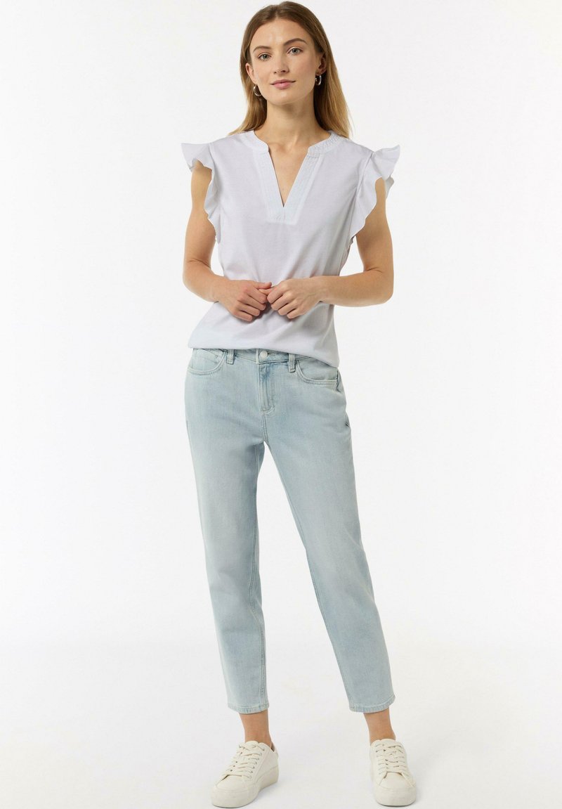 Weißes V-Ausschnitt-Top mit Rüschensäumen, kombiniert mit hellblauen, schmal zulaufenden Jeans und weißen Sneakern, stehend vor einem einfarbigen Hintergrund.