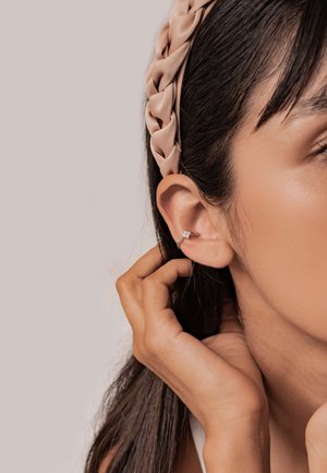 Frau, die ihr Ohr mit einer Hand berührt, trägt ein geflochtenes beiges Haarband und einen kleinen silbernen Ohrring mit einem klaren Edelstein.