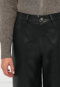 Close-up van een persoon die zwarte leren broek met een zilveren knoop en een bruine gebreide trui met rits draagt.
