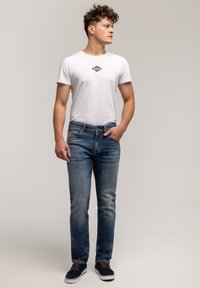 Biała koszulka z małym logo, niebieskie dżinsy i granatowe trampki z brązowymi sznurówkami. Luźny krój, klasyczny design, miękka tekstura.