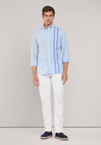 Camisa de manga larga azul clara con acentos a rayas, cuello abotonado, bolsillo en el pecho, combinada con pantalones blancos y zapatos navy.