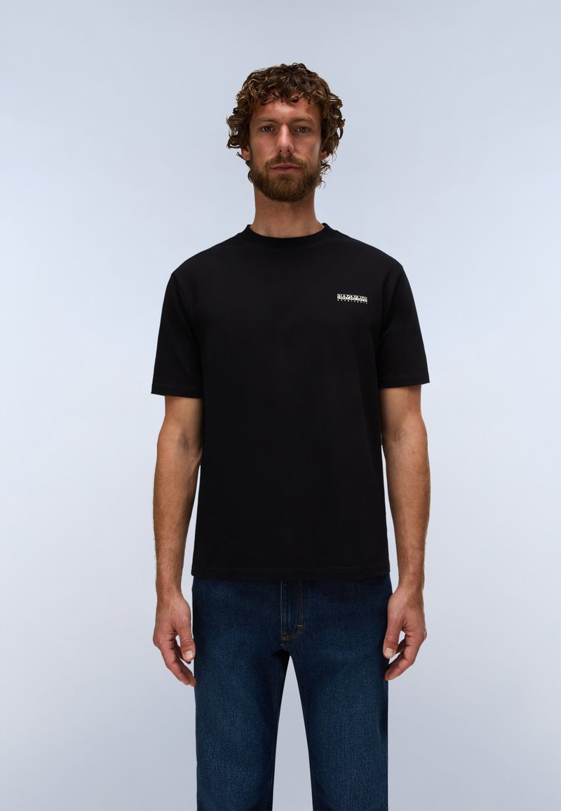 Mann mit lockigem Haar und Bart, trägt ein schwarzes Kurzarm-T-Shirt und blaue Jeans, steht vor einem schlichten, hellen Hintergrund.