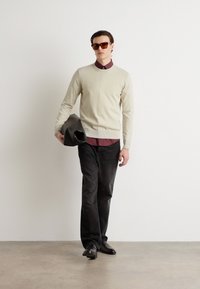 Bež pleten pulover, maroon srajca s kragnico pod njim, temni hlače in črne čevlje. Model drži črno torbico za ovojnice, nosi rjave sončne očala.