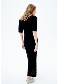 Schwarzes Strickkleid mit passgenauer Silhouette, kurzen Ärmeln, Rautenmuster und seitlichem Schlitz, kombiniert mit hellen Wedge-Schuhen.