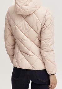 Persona che indossa una giacca a hood leggera beige trapuntata e jeans blu scuro, mostrata di spalle su uno sfondo neutro.