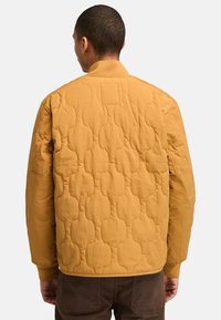 Gequilted jasje in mosterdgeel, met een gestructureerd patroon, ribgebreide kraag en mouwen. Een glad materiaal met een ontspannen pasvorm.