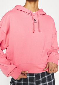 Różowa krótka bluza z kapturem z troczkiem, z małym czarnym logo Adidas. Materiał wydaje się miękki i teksturowany. Noszona na spodniach w kratę.