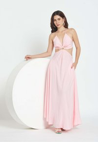 Mujer con un vestido fluido de color rosa pálido con aberturas y tirantes finos, de pie con un brazo apoyado en un gran accesorio circular blanco.