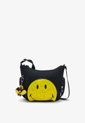 Sort crossbody-taske med stort gult smiley-ansigt-patch og lille gul abe-nøglering fastgjort til lynlås.
