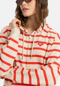 Sudadera beige con cremallera y rayas horizontales rojas, que presenta un pequeño logo rojo en el pecho y capucha con cordón. Textura de tela suave.