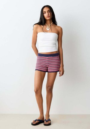 Femme aux longs cheveux foncés portant un haut blanc sans bretelles, un short en maille rayé rouge, blanc et bleu, et des tongs noires, debout contre un mur uni.