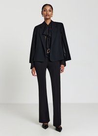 Schwarzer maßgefertigter Blazer mit ausgestellten Ärmeln, getragen über einer schwarzen Bluse mit Schleifendetail, kombiniert mit schmal geschnittenen schwarzen Hosen und spitzen Flats.