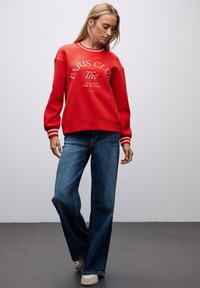 Rode sweatshirt met witte tekst "PARIS CLUB" en gestreepte afwerking, gecombineerd met blauwe wijde jeans en beige sneakers, tegen een neutrale achtergrond.