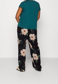 Haut teal à manches courtes avec une bordure en dentelle, associé à un pantalon large noir orné d'un grand motif floral beige et d'accents verts. Sandales noires.