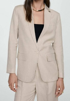 Beige Nadelstreifen-Blazer und passende Hose, getragen über einem schwarzen Oberteil, accessoriert mit einer goldenen Halskette und Ringen.