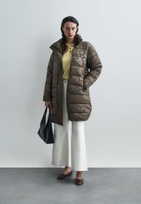 Brun pufferjakke med en skinnende tekstur, med høy krage og geometrisk mønster. Brukt over en gul topp og hvite vide bukser.