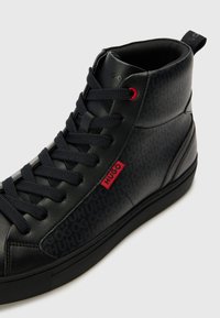 Baskets montantes noires en cuir lisse avec un motif de logo texturé, des lacets foncés, des accents rouges et une semelle en caoutchouc flexible.