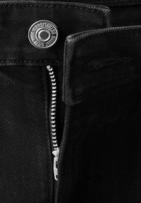 Gros plan sur un jean denim noir avec une fermeture éclair argentée partiellement ouverte et un bouton en métal portant l'inscription "MNG JEANS ESTABLISHED 1982."