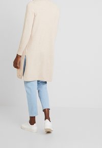 Long beige cardigan made of soft material, featuring a side slit design, worn over light blue jeans and white sneakers.